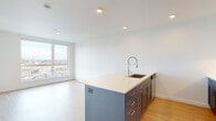 This contemporary single-floor unit showcases a clean, modern design defined by crisp white walls, light hardwood-style flooring, and an abundance of natural light. The open-concept kitchen is a standout, centered around a large island with a quartz countertop, an undermount sink with a professional-style pull-down faucet, and bold navy-blue cabinetry with brushed hardware. Full-height white upper cabinets, a subway tile backsplash, and stainless steel appliances — including a refrigerator, range, and microwave — complete the well-appointed cooking space. The living and dining areas flow seamlessly from the kitchen, with large windows framing expansive urban views of the surrounding cityscape. The bedrooms are bright and spacious, each featuring oversized windows that flood the rooms with natural light and offer elevated views of the neighborhood below. The bathroom maintains the unit's cohesive aesthetic, featuring a navy vanity with gold-toned hardware, a white countertop, a rectangular mirror, and wood-look flooring. Recessed lighting throughout adds a refined touch, while the consistent use of light tones and modern fixtures creates a bright, airy ambiance across the entire space.