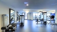 This fitness center offers a thoughtfully designed workout space finished with a smooth, dark rubberized floor that provides both durability and comfort underfoot. The layout is open and spacious, allowing for easy movement between equipment stations without feeling cramped. A collection of strength-training machines lines one side of the room, complemented by a large wall mirror that enhances the sense of depth and supports proper form during exercise. On the opposite end, cardio equipment including elliptical trainers and stationary bikes is positioned near a bank of large windows, which flood the area with natural light and offer outdoor views — creating an energizing atmosphere for workouts. The ceiling features recessed and pendant lighting that ensures bright, even illumination throughout the space. Neutral-toned walls and a clean, minimalist aesthetic contribute to the room's modern, professional feel. A glass-paneled entry door adds a polished touch to the entrance. Overall, the fitness center is well-organized and equipped to support a range of exercise routines in a clean, contemporary environment.
