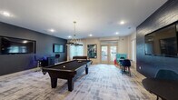 Located at 9015 Element Dr in Garner, NC, this single-story amenity space is designed to serve as a dynamic hub for recreation and productivity. The layout is thoughtfully divided into distinct zones that flow seamlessly into one another across patterned, abstract-print carpet flooring that anchors the entire space.

The game room area is a standout feature, centered around a full-size pool table with a sleek black felt surface, illuminated by a modern brass-and-black pendant chandelier overhead. A textured dark accent wall provides a dramatic backdrop for a wall-mounted flat-screen television and built-in bar-height seating. A foosball table, a connect-four game station, and additional high-top seating round out the entertainment offerings. French doors along one wall invite natural light and provide direct access to an outdoor area.

Transitioning into the coworking zone, the design shifts to a lighter, more functional palette. Oversized white dome pendant lights hang above a long communal wood-top table with industrial-style stools. Built-in white cabinetry with granite-look countertops, integrated workstations, and a computer terminal offer practical workspace amenities. A glass-paneled private meeting room with a mounted display screen provides a more enclosed setting for focused work or small gatherings.