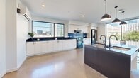 Located at 622–626 Third Ave S in Nashville, TN, this contemporary kitchen space offers a thoughtfully designed, single-floor layout that maximizes both functionality and style. The open-concept kitchen is defined by clean lines and a modern aesthetic, anchored by crisp white flat-panel cabinetry with black hardware that runs along the full length of one wall, providing generous storage. Dark, matte countertops create a striking contrast against the white cabinetry, while a bold blue vertical subway tile backsplash adds a vibrant pop of color behind the range. A central island with matching dark countertops serves as the focal point, complete with an integrated sink, dual pendant lights overhead, and bar-height seating for casual dining or gathering. Full-size stainless steel appliances, including a range with hood ventilation and a refrigerator, complete the well-equipped cooking area. The polished concrete floors lend an industrial-chic quality to the space, reflecting natural light that floods in through large, floor-to-ceiling windows. These expansive windows frame views of the surrounding Nashville skyline and greenery, bringing the outdoors in. A wall-mounted display screen and a barn-style sliding door add further contemporary touches to this stylish, versatile space.