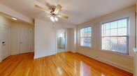This single-floor unit offers a well-organized layout that balances comfort and functionality. The main living areas are defined by gleaming hardwood floors that run throughout, lending warmth and a classic character to the space. Ceiling fans with integrated light fixtures are present in the primary rooms, providing both practical airflow and ambient lighting. Multiple windows fitted with horizontal blinds allow natural light to flow freely, creating a bright and airy atmosphere throughout the day.

The bedroom area is generously proportioned, featuring a corner window arrangement that maximizes natural light and offers views of the surrounding exterior. A radiator unit with a decorative cover adds a traditional architectural touch to this room.

The kitchen is equipped with light wood cabinetry, dark granite-style countertops, a stainless steel sink, a white refrigerator, and a gas range, all set atop clean ceramic tile flooring. A white subway tile backsplash completes the functional and tidy aesthetic of the space.

The bathroom retains a classic charm with its black-and-white mosaic tile flooring, white subway wall tiles accented by a dark border, a pedestal-style toilet, a vanity with overhead cabinetry, and a window that provides natural ventilation. Throughout the unit, clean white walls and well-maintained trim contribute to a cohesive, understated aesthetic.