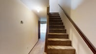 This two-story residential space offers a straightforward and functional layout spread across two well-connected floors. The interior is finished with wall-to-wall carpeting throughout, providing a warm and comfortable underfoot feel that carries consistently from room to room and along the staircase.

A central staircase serves as the primary connection between the two floors, featuring carpet-covered treads and a wooden handrail that runs the full length of the flight. The staircase is flanked by smooth, neutral-toned walls that contribute to a clean and understated aesthetic. Overhead, a flush-mount ceiling light provides focused illumination in the stairwell area.

At the base of the stairs, a hallway leads toward an adjoining room, which benefits from natural light filtering through vertical blinds — suggesting a window or sliding door that faces outward. This detail adds a sense of openness and connectivity to the exterior.

The overall ambiance of the space is modest and residential in character, with neutral wall tones and consistent carpeting creating a cohesive interior palette throughout both levels. The two-floor configuration provides a natural separation between living areas, making the space well-suited for everyday residential use.