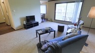 This single-floor residence features an open-concept layout that seamlessly connects the living and dining areas. The living room is carpeted and generously proportioned, accommodating a full sectional sofa, a dark-finished coffee table, and an ottoman, with ample room to move freely. A low-profile media console holds a flat-screen television along one wall, keeping the arrangement practical and uncluttered.

Natural light is a defining feature of the space, delivered primarily through a large sliding glass door that spans much of one wall. This door opens to an outdoor balcony furnished with seating, offering views of a green, landscaped exterior. A secondary window near the dining area further brightens the space.

The dining area, visible from the living room, features hardwood-style flooring that distinguishes it from the carpeted living zone, creating a subtle but functional separation. It comfortably fits a small dining table and chairs.

The overall ambiance is bright, airy, and functional, with neutral wall tones and natural light contributing to an open, welcoming feel well-suited for everyday living.