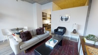 This single-floor residence showcases a thoughtfully designed open-concept layout that blends industrial and contemporary aesthetics. The living area is anchored by a comfortable grey sofa and a sleek black coffee table, set atop a richly toned area rug that adds warmth and depth to the space. A mid-century modern accent chair provides additional seating, while wall art and curated décor contribute to the room's artistic character.

One of the most striking architectural features is the exposed concrete ceiling, which transitions into a bold gold-painted section, adding a dramatic and unique visual element to the space. Structural columns punctuate the layout, reinforcing the industrial feel while defining zones within the open floor plan.

The kitchen is visible from the living area through an open passageway, offering dark cabinetry, stainless steel appliances, and open shelving — a functional and well-appointed cooking space. Wood-toned flooring runs throughout, tying the living and kitchen areas together cohesively.

The overall ambiance is modern and urban, with a curated mix of textures and materials — from raw concrete to warm metallics — creating a space that feels both sophisticated and inviting.