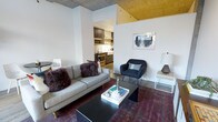 This single-floor residence showcases a thoughtfully designed open-concept layout that blends industrial and contemporary aesthetics. The living area is anchored by a comfortable grey sofa and a sleek black coffee table, set atop a richly toned area rug that adds warmth and depth to the space. A mid-century modern accent chair provides additional seating, while wall art and curated décor contribute to the room's artistic character.

One of the most striking architectural features is the exposed concrete ceiling, which transitions into a bold gold-painted section, adding a dramatic and unique visual element to the space. Structural columns punctuate the layout, reinforcing the industrial feel while defining zones within the open floor plan.

The kitchen is visible from the living area through an open passageway, offering dark cabinetry, stainless steel appliances, and open shelving — a functional and well-appointed cooking space. Wood-toned flooring runs throughout, tying the living and kitchen areas together cohesively.

The overall ambiance is modern and urban, with a curated mix of textures and materials — from raw concrete to warm metallics — creating a space that feels both sophisticated and inviting.