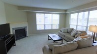 This single-floor residence features a spacious living area finished with soft, neutral-toned carpeting and warm beige walls that contribute to a relaxed and cohesive aesthetic. The room is anchored by a tile-surround fireplace set into the left wall, providing a functional and visually grounding focal point. Large windows span a significant portion of the exterior wall, allowing abundant natural light to fill the space while offering views of the surrounding neighborhood and greenery. The window arrangement creates an open, airy feel throughout the living area. Furnishings include a pair of neutral upholstered sofas arranged around a dark rectangular coffee table, complementing the room's understated color palette. The layout provides generous circulation space, making the room feel well-proportioned and comfortable for everyday living. Recessed ceiling details add a subtle architectural element, while the overall design emphasizes simplicity and functionality. The combination of natural light, soft textures, and a neutral palette gives the space a calm, welcoming ambiance suitable for both relaxing and entertaining.