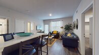 This single-floor unit offers a thoughtfully designed layout that balances communal living with private spaces. The entryway welcomes residents with a clean, well-lit hallway finished with wood-look flooring, providing a seamless transition into the main living areas.

The open-concept living and dining area is a central highlight of the unit, featuring large windows dressed with horizontal blinds that invite generous natural light. The living room accommodates a comfortable seating arrangement and includes a wall-mounted television, an exterior door, and a relaxed, welcoming atmosphere.

The kitchen is outfitted with dark, modern cabinetry paired with light countertops, stainless steel appliances, and ample storage. A raised bar counter with seating connects the kitchen to the dining area, making it ideal for both meal preparation and casual dining.

The unit includes multiple private bedrooms, each featuring wood-look flooring, a window for natural light, built-in closet shelving, and space for a bed and desk — well-suited for study and rest. Two full bathrooms serve the unit, each equipped with a tub-shower combination featuring tiled surrounds with a contrasting dark accent band, a single-sink vanity with dark cabinetry, and wood-look tile flooring. The overall aesthetic is modern and cohesive throughout.