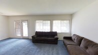This single-floor space centers around a generously sized living room that provides a comfortable and practical setting. The room is finished with blue-grey carpeting that extends throughout the area, adding a soft, casual feel underfoot. White walls and a smooth, neutral ceiling contribute to a clean, understated aesthetic.

Natural light is a notable feature of this living space, delivered through a row of large windows fitted with horizontal blinds, allowing occupants to control the level of light and privacy as needed. The windows face outward toward a treed exterior, providing a pleasant outdoor view. A glazed door on the left wall offers direct access to the outside, further enhancing the connection to the outdoors and allowing additional natural light into the room.

The layout of the living area comfortably accommodates multiple seating pieces, as evidenced by the placement of two dark upholstered sofas arranged to make use of the available floor space. The room's proportions allow for flexible furniture arrangements, making it well-suited for everyday living or entertaining. Overall, the space presents a functional, light-filled environment with a straightforward, livable character.