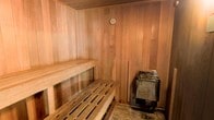 This traditional sauna is a dedicated wellness retreat, constructed entirely with warm, natural cedar wood that lines every wall, ceiling, and floor surface. The rich, honey-toned wood paneling creates an inviting and calming atmosphere, characteristic of a well-crafted sauna environment. The interior is thoughtfully laid out with two tiered wooden benches — an upper and lower level — allowing users to choose their preferred level of heat intensity, as temperatures naturally rise toward the ceiling. A stainless steel electric sauna heater is positioned in the corner, serving as the heart of the space and efficiently generating the dry heat essential for an authentic sauna session. A round thermometer is mounted on the upper wall, enabling precise monitoring of the interior temperature. The natural wood construction not only contributes to the aesthetic warmth of the space but also plays a functional role in absorbing and radiating heat evenly throughout the room. The overall design is clean, minimalist, and purposeful, prioritizing comfort and function. This sauna offers an intimate and restorative environment suited for relaxation, muscle recovery, and overall well-being.