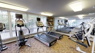 This fitness center offers a comprehensive workout environment spread across a single floor, thoughtfully divided into distinct exercise zones. The cardio area is outfitted with a row of Star Trac treadmills equipped with individual display screens, positioned in front of expansive floor-to-ceiling windows that frame views of surrounding greenery and fill the room with natural light. A large wall mirror enhances the sense of space and allows users to monitor their form during exercise. Flush-mounted rectangular lighting fixtures supplement the natural illumination, creating a bright and energizing atmosphere.

Adjacent to the cardio zone, a generously proportioned strength training area features a variety of cable and weight machines with ergonomic padding, catering to a full range of muscle groups. An adjustable workout bench adds versatility for free-weight exercises. The room is finished with textured wallcovering, patterned carpeting, and elegant ceiling fixtures that lend a classic, well-maintained character to the space.

A wide connecting corridor links the two exercise areas and provides additional open floor space, complete with a wall-mounted fire safety station and a water fountain, ensuring both safety and convenience throughout the facility.
