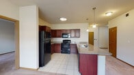 This single-floor unit offers a well-designed, open-concept layout that seamlessly connects the kitchen, dining, and living areas. The kitchen is outfitted with rich, dark cherry wood cabinetry offering ample storage, granite-style countertops, black appliances including a refrigerator, electric range, and built-in microwave, and a large peninsula with a double sink — finished with elegant pendant lighting overhead. Light tile flooring runs throughout the kitchen, transitioning to plush carpeting in the living and bedroom areas.

The spacious living area is bathed in natural light from a bank of large windows, and features a ceiling fan for added comfort. A wide, open hallway connects the main living spaces to the private rooms, with arched interior doors adding a refined architectural touch throughout.

The primary bedroom is generously sized with soft carpeting, a ceiling fan, and a window with blinds. It connects directly to a bathroom that showcases a distinctive dark wood vanity with ornate detailing, a black countertop, a framed decorative mirror, tile flooring, and access to a walk-in closet. An additional bedroom provides comfortable proportions with carpeting, a ceiling fan, and natural light. The layout is practical, polished, and move-in ready.