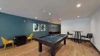 This single-floor amenity space is a well-appointed game and social lounge designed for recreation and community gathering. The layout is generously proportioned, featuring warm-toned hardwood flooring throughout that lends a cohesive and inviting feel to the entire space.

At the heart of the room sits a full-size pool table with a slate-blue felt surface, positioned beneath pendant lighting that adds a focused, atmospheric glow. Adjacent to it, a shuffleboard table extends along one wall, accompanied by vibrant abstract artwork that adds pops of color against the teal accent walls. A dedicated media area features a large wall-mounted flat-screen television framed by textured acoustic paneling, creating an immersive viewing experience. Bright yellow accent chairs and a compact game table nearby provide casual seating for social interaction.

The space also includes a standing-height bar table with seating, open shelving units, and additional flexible seating arrangements throughout. Recessed lighting illuminates the room uniformly from above.

The entrance foyer, branded with a "Nolan" floor mat, connects seamlessly to the lounge and benefits from floor-to-ceiling glass doors that flood the space with natural light. Large sliding glass doors at the far end open directly to an outdoor patio courtyard, further extending the social environment and providing easy indoor-outdoor access.