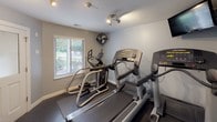 This single-level fitness center offers a practical and functional workout environment designed to meet a variety of exercise needs. The space is finished with light gray walls, rubber flooring, and recessed and track lighting that keeps the area well-lit throughout. A wall-mounted flat-screen television is positioned near the cardio equipment, providing entertainment during workouts.

The main exercise area houses multiple Life Fitness treadmills and at least one elliptical cross-trainer, arranged to allow comfortable use of each machine. A wall-mounted fan provides additional air circulation. A window overlooking a landscaped outdoor area allows natural light to enter and offers a pleasant view while exercising.

A dedicated free weights section features a tiered dumbbell rack stocked with a range of weights, accompanied by a wall mirror that enhances the sense of space and supports proper form during strength training. A frosted-glass partition separates an additional exercise room, visible through the entryway. Safety equipment including a fire extinguisher and alarm are present, along with posted facility rules and hours of operation. The overall ambiance is clean, organized, and purposefully utilitarian.