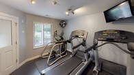 This single-level fitness center offers a practical and functional workout environment designed to meet a variety of exercise needs. The space is finished with light gray walls, rubber flooring, and recessed and track lighting that keeps the area well-lit throughout. A wall-mounted flat-screen television is positioned near the cardio equipment, providing entertainment during workouts.

The main exercise area houses multiple Life Fitness treadmills and at least one elliptical cross-trainer, arranged to allow comfortable use of each machine. A wall-mounted fan provides additional air circulation. A window overlooking a landscaped outdoor area allows natural light to enter and offers a pleasant view while exercising.

A dedicated free weights section features a tiered dumbbell rack stocked with a range of weights, accompanied by a wall mirror that enhances the sense of space and supports proper form during strength training. A frosted-glass partition separates an additional exercise room, visible through the entryway. Safety equipment including a fire extinguisher and alarm are present, along with posted facility rules and hours of operation. The overall ambiance is clean, organized, and purposefully utilitarian.