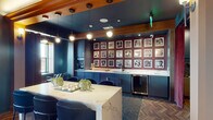 This single-floor space is designed with a sophisticated, eclectic aesthetic that combines bold color choices, varied textures, and carefully curated furnishings. The layout flows between several distinct functional zones unified by a cohesive design language.

The wet bar and kitchen area serves as a central focal point, featuring deep navy cabinetry paired with a dramatic white marble-veined countertop island. Black bar stools line the island, while a grid of black-and-white framed photographs mounted against a burgundy accent wall creates a gallery-like backdrop. A built-in beverage refrigerator and pendant globe lighting complete the space. Flooring in this zone transitions between hexagonal dark tile and herringbone wood parquet.

The adjoining lounge area is anchored by a tufted brown leather sectional sofa, vintage-style egg chairs in cognac leather, and a teal accent chair atop layered Persian-style rugs. A fireplace with an integrated flat-screen television serves as the focal point, while deep teal walls and warm cove lighting above wood beam detailing add depth and intimacy.

A corridor connects the zones, featuring the same herringbone flooring and globe pendant fixtures. French doors open onto an outdoor terrace with seating and panoramic city views, allowing natural light to flood the interior and providing a seamless indoor-outdoor connection.