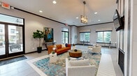Located at 7145 Anderson Dr in Whitestown, Indiana, this well-appointed single-floor clubhouse offers a thoughtfully designed communal space ideal for gathering and relaxation. The open-concept lounge area serves as the heart of the space, featuring light-toned large-format tile flooring, recessed lighting, and a geometric chandelier that anchors the seating area. A bold tufted cognac leather sofa, neutral upholstered chairs, and a colorful abstract area rug create an inviting, curated atmosphere. Large windows along the perimeter flood the interior with natural light and provide views of the surrounding greenery. A textured, pebble-stone accent wall frames a wall-mounted flat-screen TV and a sleek built-in fireplace, adding warmth and visual interest to the space. The adjacent kitchen is equally impressive, showcasing rich mahogany cabinetry, dark granite countertops, a central island with bar seating, and a striking cage-style chandelier overhead. A beverage refrigerator, microwave, and dedicated refreshment station make this kitchen fully functional for hosting. Accent walls in deep navy blue provide bold contrast throughout, while the overall design strikes a balance between modern sophistication and comfortable functionality.