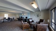 This single-floor space is thoughtfully designed to serve both productivity and relaxation, combining a functional business center with a stylish lounge area. The workstation zone is equipped with multiple desktop computers arranged along a dedicated desk, paired with ergonomic rolling chairs — ideal for focused work sessions. Overhead, a bold fluorescent fixture provides ample task lighting to keep the area bright and functional.

Transitioning seamlessly into the lounge area, rich brown leather armchairs are arranged around a round glass-top accent table, creating an inviting setting for casual meetings or unwinding. Warm pendant lights hang above, casting a softer glow that complements the cooler tones of the adjacent work zone. The walls feature a soothing blue-gray accent color that adds depth and a contemporary feel to the space.

Decorative touches, including a starburst mirror wall sculpture and framed artwork, lend the room a mid-century modern aesthetic. The exposed structural beams on the ceiling add an industrial character, while the patterned carpet grounds the space with texture and warmth. Overall, the space strikes a well-balanced blend of professional utility and comfortable design, making it well-suited for both work and leisure.