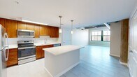 This one-bedroom, one-bathroom unit showcases a compelling mix of industrial loft character and contemporary design across a single floor. The open-concept living and kitchen area forms the heart of the space, where light-toned large-format tile flooring extends throughout, creating a cohesive and airy feel. The kitchen is well-appointed with warm-toned wood cabinetry, stainless steel appliances including a refrigerator, range, microwave, and dishwasher, and a light-colored countertop that continues onto a central island. Pendant lighting above the island adds a refined touch, while recessed and under-cabinet lighting enhances functionality.

The living area opens toward large windows that frame outdoor city views, allowing natural light to fill the space. Exposed concrete beams, visible ductwork, and raw structural elements on the ceiling lend an authentic loft aesthetic that contrasts attractively with the clean white walls. The bedroom is a comfortable size, featuring the same large-format tile flooring, tall windows with city views, and the signature exposed beam ceiling. The bathroom is finished with tile flooring, a wood-toned vanity, white countertop, and a full soaking tub with subway tile surround.