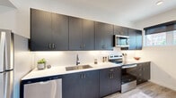 This thoughtfully designed single-floor unit offers a streamlined, modern aesthetic throughout. The open-concept living and dining area is anchored by light wood-look flooring, recessed lighting, and crisp white walls that create a bright, airy atmosphere. A sliding glass door leads to a private balcony, offering outdoor views and allowing natural light to flow generously into the space.

The kitchen is a standout feature, finished with dark charcoal cabinetry accented by brushed gold hardware, white quartz countertops, and a classic subway tile backsplash. Stainless steel appliances — including a range, microwave, and dishwasher — complete the functional and stylish cooking space.

The bedroom is a calm, well-proportioned retreat with neutral tones, carpeted flooring, and a window that brings in soft natural light. The bathroom is clean and contemporary, featuring a combined shower and tub, a dark vanity with gold hardware, a backlit mirror, and wood-look tile flooring. Notably, a stacked in-unit washer and dryer is incorporated directly into the bathroom for added convenience.

The entryway is clean and minimal, with a keypad entry door and a wall-mounted media console that doubles as a functional accent piece. Overall, the space delivers a cohesive modern design with practical, high-quality finishes throughout.