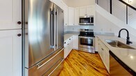 Located at 18 W Main St in Hornell, NY, this two-story loft-style space masterfully combines industrial heritage with contemporary design. The main level showcases a sweeping open-plan living area anchored by warm, reclaimed hardwood floors that contrast beautifully against exposed painted brick walls. Soaring ceilings are accented by dark structural beams and an exposed ductwork system, lending an authentic industrial aesthetic. Oversized arched windows dressed with roller shades flood the space with natural light, while ceiling fans and pendant lighting add both function and style. A modern kitchen flows seamlessly from the main living area, outfitted with white shaker cabinetry, subway tile backsplash, grey countertops, a large island, and a full suite of stainless steel appliances. A sleek staircase with black metal railings leads to the upper level, where the architecture takes on a dramatic character — exposed dark rafters create angled, vaulted rooflines that define the upper rooms. A well-appointed bathroom on this level features large-format tile, a frameless glass sliding shower enclosure, and brushed nickel grab bars and fixtures for both style and accessibility. The overall ambiance blends raw, historic charm with thoughtfully curated modern upgrades throughout.