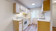 This single-floor unit presents a clean, functional layout with a straightforward flow between rooms. The kitchen is designed for efficiency, featuring a galley-style configuration with warm wood-toned lower cabinetry paired with white upper cabinets, light countertops, and white appliances including an electric range, refrigerator, and dishwasher. Wood-look laminate flooring runs through the kitchen and hallway areas, adding warmth and easy maintenance. A vertical blind-covered window provides natural light to the kitchen.

The hallway connects the main living areas with a sliding door closet for additional storage. Bedrooms are carpeted in neutral tones and offer generous dimensions. One bedroom features dual closets with built-in shelving and hanging rods, providing substantial storage capacity, along with a ceiling fan with light fixture. A second bedroom offers a window with natural light and similarly neutral carpeting and walls.

The bathroom is finished with wood-look flooring, a white vanity with an integrated sink, a large wall mirror, and a full tub-shower combination with white tile surround. Throughout the unit, white walls and flush-mount ceiling lighting create a bright, clean aesthetic.
