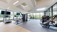 Located at 1600 James Naismith Drive in Ottawa, Ontario, this modern fitness facility offers a thoughtfully designed, single-floor space that combines functionality with an bright, open atmosphere. The gym is bathed in natural light courtesy of expansive floor-to-ceiling windows on multiple walls and large skylights integrated into the coffered ceiling, providing sweeping views of the surrounding greenery.

The main workout area features rubber flooring across a generously sized exercise zone, housing a comprehensive selection of equipment including treadmills, cable machines, a power rack with weight storage, benches, dumbbells, and stationary and recumbent bikes. Light hardwood flooring accents the perimeter areas, adding warmth to the space.

A bold orange accent wall featuring motivational branding adds a dynamic visual element, while black-framed glass partitions divide the space into distinct zones, including a separate wellness or activity room visible through the glass. The facility also includes a water fountain station for convenience.

The overall ambiance is energetic yet refined, with high ceilings and abundant natural light creating an inviting environment. Outdoor terrace views and access to surrounding landscaped areas further enhance the experience, making this a well-appointed fitness destination within the broader complex.