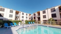 This outdoor pool amenity area is set within a multi-story residential complex, offering a welcoming retreat for residents. The centerpiece is a generously sized swimming pool with clear, blue water, complemented by an adjacent spa, providing options for both active swimming and relaxed soaking. The surrounding concrete sun deck is spacious, furnished with a variety of lounge chairs and chaise lounges positioned to take full advantage of the open sky and natural sunlight.

The pool area is enclosed by a metal perimeter fence, ensuring a secure and private environment. Lush greenery and mature trees line portions of the space, adding natural shade and visual appeal. Multiple residential buildings frame the area, featuring balconies and large windows that overlook the pool courtyard.

Access to the pool is provided through a dedicated entrance point with a door framed by wood trim and equipped with wall-mounted lighting. A pool rules sign is posted near the entrance, indicating a managed and well-maintained facility. A small covered transitional space adjacent to the entrance allows residents to comfortably move between the interior and the pool deck. The overall ambiance is bright, open, and relaxed, making this pool area a functional and attractive communal outdoor space.