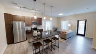 This 2-bedroom, 2-bathroom residence is thoughtfully designed to maximize comfort and functionality across a single-floor layout. The heart of the home is an open-concept kitchen and living area that seamlessly connects the two spaces, creating an inviting and airy atmosphere. The kitchen is outfitted with dark granite countertops, rich dark-wood cabinetry, and stainless steel appliances, including a refrigerator, microwave, and range. A large central island with an extended breakfast bar and industrial-style stools serves as both a prep space and a casual dining area. Pendant lighting hangs above the island, complementing the track lighting overhead. Light wood-style flooring runs throughout the main living area, adding warmth and continuity. The adjacent living room benefits from multiple windows dressed with sheer curtains, allowing generous natural light to fill the space and creating a bright, welcoming ambiance. The two bedrooms provide comfortable private retreats, each supported by a full bathroom. The overall design balances modern aesthetics with practical living, featuring clean lines, neutral tones, and contemporary finishes that give the home a polished and move-in-ready feel.