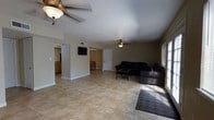 This single-story space presents an open-concept layout that connects the living area to the kitchen, creating a functional and cohesive floor plan. The living room is generously sized, featuring large neutral-toned ceramic tile flooring that extends throughout the space, contributing to a clean and consistent aesthetic. Two ceiling fans with light fixtures are installed overhead, providing both ambient lighting and air circulation.

The living area accommodates a seating arrangement centered around a low coffee table, with a wall-mounted flat-screen television positioned for comfortable viewing. Natural light enters through a window on one side, adding brightness to the room. A doorway near the television leads to additional areas of the home.

The adjacent kitchen is visible through an open passage, revealing wood-toned cabinetry and a dining area beyond. The neutral beige walls throughout maintain a simple, understated tone that complements the tile flooring. A door near the kitchen side of the living area provides access to another section of the home. Overall, the space offers a straightforward, practical layout suited for everyday living.