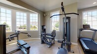 Located at 6460 Chelsea Glen Dr in Canal Winchester, Ohio, this community fitness center is a single-floor facility thoughtfully designed to support a well-rounded workout experience. The space is organized into distinct exercise zones, each outfitted with dedicated equipment to serve a variety of fitness needs.

The cardio area features a row of Bodycraft treadmills aligned beneath a bank of large, grid-paned windows that flood the room with natural light and offer pleasant views of the surrounding neighborhood. Elliptical trainers and additional treadmills are positioned nearby, also benefiting from generous window coverage and vaulted ceilings that lend an open, airy feel to the space.

A separate strength training area houses a multi-station cable and weight machine alongside a leg press and a flat bench, providing comprehensive resistance training options. This zone is equally well-lit, with multiple windows framing outdoor views.

The main open exercise floor is expansive and finished with polished concrete, making it well-suited for stretching, functional training, or group exercise. Full-length mirrors line the walls, visually expanding the space and assisting with form. Recessed lighting and vaulted ceilings enhance the bright, motivating ambiance throughout.

French doors connect the main floor to adjacent areas, including what appears to be a mailroom or community corridor, indicating this fitness center is part of a broader community amenity building.