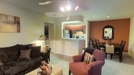 This single-story space features an open-concept layout that seamlessly connects the living area to the kitchen. The living room is carpeted and comfortably sized, accommodating a seating arrangement that includes plush armchairs and a sofa, centered around a decorative ottoman. A ceiling fan with an integrated light fixture provides both ventilation and overhead illumination. A warm terracotta accent wall adds a distinctive design element, complementing the neutral tones found throughout the rest of the space.

The kitchen is accessible via a pass-through counter that opens toward the living area, creating a functional and connected feel. It is equipped with wood-toned cabinetry, a white range, and a refrigerator. Under-cabinet lighting adds a subtle glow to the workspace. The pass-through design allows for interaction between the kitchen and living areas, making it well-suited for entertaining.

Adjacent to the living room, a dining area is visible, furnished with a dark wood table and chairs. A hallway leads to additional rooms, and a framed mirror near the entryway enhances the sense of depth. The overall ambiance is warm and inviting, with layered lighting and a cohesive color palette throughout.