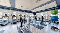 Located at 2439 Casona Ln in Melbourne, Florida, this community fitness center offers a well-equipped, single-floor workout facility designed to support a comprehensive exercise routine. The space is defined by its bold navy blue and white color scheme, with deep blue accent bands lining the upper walls and a raised tray ceiling that adds architectural depth and a sense of height to the interior. Light wood-look flooring runs throughout, complementing the clean, modern aesthetic.

Natural light floods the space through an abundance of large, multi-pane windows that wrap around the room on multiple sides, providing open views of the surrounding community and keeping the atmosphere bright and energizing. Ceiling fans and recessed lighting ensure comfortable conditions throughout the day.

The fitness floor is generously proportioned and thoughtfully organized into distinct zones. The cardio section features multiple treadmills, StairMaster steppers, elliptical trainers, stationary bikes, and a rowing machine. The strength training area is equally well-stocked, with a variety of selectorized cable machines, weight stations, and a full dumbbell rack. Exercise mats and stability balls are also available, supporting stretching and functional fitness. Wall-mounted televisions are positioned for easy viewing during workouts. Sanitation supplies and a wall clock complete the practical amenities of this polished, community-oriented fitness space.