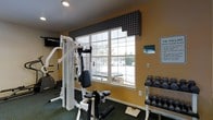 This fitness room offers a practical and well-equipped workout space designed to accommodate a variety of exercise routines. The room features warm yellow walls that create an inviting atmosphere, complemented by a dark green patterned carpet that adds a classic touch to the space.

A prominent multi-station cable and weight machine serves as the centerpiece of the room, providing options for a wide range of strength-training exercises. To the right, a organized dumbbell rack holds a full selection of free weights, catering to users of varying fitness levels. A cardio machine, visible toward the back corner, rounds out the equipment offerings.

A large window dressed with a plaid fabric valance allows natural light to fill the room and provides views of the surrounding outdoor area, enhancing the overall atmosphere during workouts. Recessed lighting in the ceiling ensures the space remains well-lit regardless of the time of day.

Posted signage on the walls provides exercise room guidelines and rules, reflecting a structured and well-managed facility. The room is thoughtfully arranged to maximize the available floor space while keeping equipment accessible and the layout functional.
