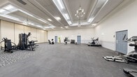 Located at 1944 Cedar Creek Rd in Fayetteville, NC, this fitness facility offers a generously sized, single-floor workout space designed to accommodate a wide range of exercise activities. The room makes a striking first impression with its soaring coffered ceilings adorned with elegant chandeliers and recessed skylights, flooding the space with bright, even light and lending an upscale ambiance uncommon in typical fitness settings.

The flooring is a practical combination of large-format rubber tiles in a neutral dark gray, providing cushioning and durability for high-impact activities, transitioning to patterned carpet in designated equipment zones. The space is equipped with a robust selection of fitness equipment, including treadmills, elliptical trainers, stationary bikes, a multi-station cable and strength machine, adjustable dumbbells on a dedicated rack, and various benches and accessories.

A standout architectural feature is the set of tall, operable room divider panels along one wall, allowing the space to be sectioned off and reconfigured as needed — ideal for group fitness classes or private training sessions. Wall-mounted mirrors near the free weight area enhance the sense of openness and support proper form during workouts. Multiple blue access doors provide convenient entry and exit points throughout the room. The clean white walls and wainscoting contribute to a bright, welcoming environment suitable for all fitness levels.