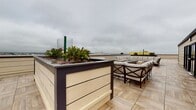 Located at 20 Burling Ln in New Rochelle, NY, this single-floor commercial space offers a functional layout with a notable rooftop terrace as a standout feature. The rooftop area is outfitted with wood-paneled raised planter beds filled with greenery, porcelain tile flooring, and a generous seating arrangement consisting of upholstered sectional and accent chairs — designed for social gatherings or relaxation with open panoramic sky views of the surrounding urban environment.

The interior portions of the space reflect a utilitarian character, with neutral off-white walls, dark baseboards, and light-toned flooring throughout. A dedicated mechanical or utility room houses exposed wiring panels, conduit runs, and infrastructure cabling mounted on a plywood backing board, indicating a space equipped for significant electrical and communications systems. A standard solid-core interior door with metal hardware provides access between areas. Ventilation is supported by wall-mounted HVAC registers. The interiors appear to be in an active construction or fit-out phase, with materials and equipment present throughout. Overall, the space combines practical infrastructure with an appealing rooftop amenity, making it well-suited for commercial or mixed-use occupancy.