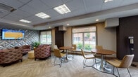 Located at 10 Kings Rd in Madison, NJ, this thoughtfully designed single-floor commercial space blends professional functionality with comfortable, contemporary aesthetics. The layout includes a well-appointed reception corridor lined with framed photography artwork, a leather bench, and a reception desk with pendant lighting — setting a polished tone from the moment of entry.

The open lounge area serves as a central gathering hub, furnished with patterned upholstered club chairs, round café-style tables, and modern tulip-base seating. A textured geometric accent wall adds visual interest, while a wall-mounted display screen provides an informational or entertainment focal point. Natural light floods the space through multiple large windows, offering views of the surrounding greenery and streetscape.

A dedicated conference room accommodates a full-sized rectangular table surrounded by cushioned executive chairs, with a window providing natural light and views of the exterior. The room features a ceiling-mounted HVAC unit and recessed lighting for a clean, professional finish.

Throughout the space, neutral-toned carpet tile flooring — including herringbone-patterned sections — anchors the design, while a drop ceiling with recessed and panel lighting ensures consistent, comfortable illumination. The overall ambiance is welcoming and versatile, well-suited for professional meetings, collaborative work, and community engagement.