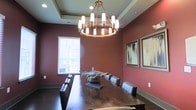 This single-story space centers around a striking dining or meeting room designed with both sophistication and visual impact. The room is defined by deep, warm red walls that create an intimate yet dramatic atmosphere, complemented by a tray ceiling that adds architectural depth and a sense of refined structure. A large, dark wood table dominates the space, surrounded by dark leather chairs that reinforce the room's bold, upscale character.

Suspended above the table is a substantial brass and white cylindrical chandelier, serving as the room's focal centerpiece. Its warm, ambient glow enhances the richness of the surrounding tones while providing functional illumination. A window along one wall allows natural light to filter into the space, softening the intensity of the wall color and offering a visual connection to the exterior.

The walls are adorned with framed abstract artwork, adding a contemporary artistic element that balances the traditional furnishings. A decorative shark sculpture displayed on the table introduces an unexpected, conversation-worthy accent piece. Together, these elements create a space that is both purposeful and visually compelling, suitable for formal dining or private gatherings.