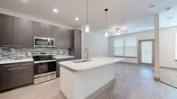 Located at 2161 Lakewood Ranch Boulevard in Sarasota, FL, this efficiently designed single-floor unit offers a thoughtfully planned open-concept layout. The kitchen serves as the centerpiece of the space, showcasing dark wood-grain cabinetry with brushed nickel hardware, crisp white quartz countertops, and a full suite of stainless steel appliances including a range, over-the-range microwave, and refrigerator. A striking mosaic tile backsplash in blues and grays adds a contemporary accent. A large white kitchen island with a built-in sink and pendant lighting above doubles as additional prep space and a casual dining area.

The open living area flows seamlessly from the kitchen, featuring light wood-style flooring, a ceiling fan, and multiple windows with white blinds that invite natural light throughout the space. A private entry door provides direct access to the unit.

The bathroom is finished in a clean, neutral palette with large-format floor tiles, a combination tub and shower with tiled surround, and a vanity with wood-tone cabinetry and a white countertop. A dedicated closet provides practical storage. The overall design aesthetic is modern and cohesive, with consistent finishes and an efficient use of space throughout.