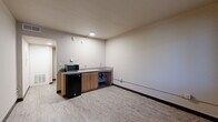 Located at 3541 E Van Buren St in Phoenix, Arizona, this compact single-floor unit offers an efficient and functional layout comprising a living room, a full bathroom, and a connecting hallway. The living area features light wood-look vinyl flooring, neutral beige walls, and a built-in kitchenette along one wall. The kitchenette includes a compact countertop with a sink, warm wood-toned cabinetry, a compact refrigerator, and a microwave, providing essential conveniences within a streamlined footprint. A window along the living area wall allows natural light to brighten the space and offers a view of the surrounding greenery outside. The room is finished with dark baseboards that provide a clean contrast to the light flooring. The bathroom is well-appointed with large-format tile flooring, a combination bathtub and shower, a toilet, and a vanity featuring wood cabinetry topped with a speckled grey countertop and an integrated sink. The hallway connects the main living space to the bathroom, offering a clear and logical flow throughout the unit. Overall, the space presents a clean, modern, and utilitarian design well-suited for single occupancy in the Phoenix area.