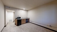 Located at 3541 E Van Buren St in Phoenix, Arizona, this compact single-floor unit offers an efficient and functional layout comprising a living room, a full bathroom, and a connecting hallway. The living area features light wood-look vinyl flooring, neutral beige walls, and a built-in kitchenette along one wall. The kitchenette includes a compact countertop with a sink, warm wood-toned cabinetry, a compact refrigerator, and a microwave, providing essential conveniences within a streamlined footprint. A window along the living area wall allows natural light to brighten the space and offers a view of the surrounding greenery outside. The room is finished with dark baseboards that provide a clean contrast to the light flooring. The bathroom is well-appointed with large-format tile flooring, a combination bathtub and shower, a toilet, and a vanity featuring wood cabinetry topped with a speckled grey countertop and an integrated sink. The hallway connects the main living space to the bathroom, offering a clear and logical flow throughout the unit. Overall, the space presents a clean, modern, and utilitarian design well-suited for single occupancy in the Phoenix area.