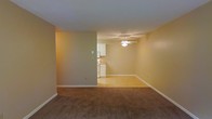 This single-floor space offers a straightforward, functional layout with a comfortable open-plan design. The living area is finished with warm, neutral-toned carpeting that adds a soft underfoot feel, while the walls are painted in a light beige hue that lends a calm, understated ambiance throughout. A ceiling fan is centrally mounted, providing both lighting and airflow to the main living space.

The living area flows naturally into a dining or transitional space, distinguished by a shift to light hardwood-style flooring that visually connects the rooms while adding a clean contrast. This area benefits from the same warm wall tones and white baseboard trim that runs consistently throughout, giving the space a cohesive, tidy appearance.

The kitchen is visible from the living area and features white cabinetry that reaches toward the ceiling, offering ample storage. The countertops provide functional workspace, and the kitchen's light flooring complements the cabinetry for a bright, clean look. Standard appliances are included within the kitchen space.

Overall, the unit presents a practical and well-organized layout with consistent finishes, neutral tones, and a seamless transition between the living and kitchen areas, making it a functional and move-in ready space.