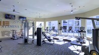 This single-floor fitness center is designed to support a comprehensive workout experience. The space is outfitted with a row of treadmills and upright cardio machines positioned along the perimeter, taking full advantage of the large windows that line the walls and flood the room with natural light. Views of the surrounding building exterior are visible through the glass, creating an open, connected feel. A cable and pulley strength training system anchors one side of the room, accompanied by adjustable benches and additional free-standing equipment. The flooring features a patterned, multi-tonal carpet that defines the workout zones while adding visual interest. Overhead, ceiling fans are suspended from a smooth, light-colored ceiling fitted with recessed lighting, ensuring comfortable air circulation and even illumination throughout the day. A motivational mural with the word "AGILITY" is painted on one wall, reinforcing the energetic atmosphere of the space. Mounted monitors are positioned near the cardio equipment for entertainment during workouts. The overall layout is organized and spacious, allowing users to move freely between stations.