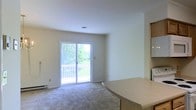This single-floor space presents a clean, functional layout centered around a bright and open living area. The room is finished with neutral beige carpeting and soft white walls, creating a calm, understated atmosphere throughout. A prominent sliding glass door serves as the focal point of the main living area, allowing abundant natural light to fill the space while framing pleasant views of lush green trees and an adjacent outdoor deck with a white railing. This seamless indoor-outdoor connection enhances the sense of openness and provides easy access to the exterior. A baseboard heating unit runs along the perimeter wall, indicating a practical, efficient heating system. To one side, a partial view of light wood cabinetry suggests a kitchen or dining area nearby, hinting at a connected, open-concept design between the living and kitchen spaces. The overall layout is straightforward and versatile, well-suited for comfortable everyday living. The neutral color palette and soft natural light give the space a welcoming, airy quality, while the surrounding greenery visible through the glass door adds a quiet, nature-connected ambiance to the interior.