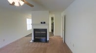 This two-story home offers a straightforward and functional layout designed for comfortable everyday living. The main living area serves as the heart of the home, featuring a classic wood-mantled fireplace with a dark surround that acts as a natural focal point in the room. Soft carpet flooring extends throughout the space, adding warmth underfoot, while a ceiling fan overhead provides efficient air circulation. Natural light filters in through a window dressed with horizontal blinds, keeping the space bright and inviting. The neutral, light-toned walls throughout create a clean and versatile backdrop that complements a variety of interior styles. An open sightline from the living area leads toward an adjacent room, hinting at a connected and accessible floor plan. A doorway visible from the main space provides easy access to additional rooms, while a glimpse into what appears to be a kitchen with darker cabinetry adds functional depth to the layout. The two-story configuration of the home provides additional living or sleeping space on the upper level, making this a well-rounded property suited for a range of occupants.