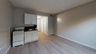 Located at 15001 Crenshaw Blvd in Gardena, California, this single-floor unit offers a practical and well-organized layout across four distinct spaces: a living room, bathroom, hallway, and closet. The living room serves as the primary multipurpose area, featuring light-toned hardwood-style flooring, neutral gray walls, and a wall-mounted air conditioning unit beneath a window that allows natural light to filter in. A compact kitchen is integrated into the living area, equipped with white cabinetry, a gas range, and a sink with dark countertops — providing efficient functionality within a modest footprint. The hallway connects the main living space to the entry door and features a textured ceiling, wall sconce lighting, and a sliding closet door for convenient storage access. A utility panel is also visible along the hallway wall. The bathroom is finished with floor-to-ceiling dark earth-toned tile surrounding a full soaking tub and shower combination, complemented by a white vanity and toilet. The tile detailing adds a bold, contrasting design element to the otherwise light and understated palette of the unit. Throughout the space, clean white baseboards and consistent flooring tie the rooms together cohesively, creating a functional and straightforward living environment.