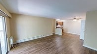 This single-floor space offers a clean, functional layout centered around an open-concept living and kitchen area. The main living space is generously sized, featuring durable hardwood-style plank flooring that runs throughout and provides a warm, cohesive aesthetic. Neutral, light-toned walls keep the interior bright and versatile, ready to complement a variety of décor styles.

A baseboard heating unit runs along the exterior wall of the living area, ensuring efficient and consistent warmth during colder months. The space transitions seamlessly into the kitchen, which is defined by a raised peninsula counter — ideal for casual dining or additional prep space. The kitchen is equipped with warm-toned wood cabinetry that offers ample storage, and the darker countertops provide a practical contrast. Overhead lighting illuminates the kitchen workspace effectively.

Toward the far end of the space, a ceiling fan adds both functionality and a finishing touch, supporting air circulation throughout the open floor plan. The overall layout is straightforward and efficient, making excellent use of the available square footage. The combination of open sightlines, consistent flooring, and a well-integrated kitchen makes this a practical and comfortable living environment suited for everyday use.