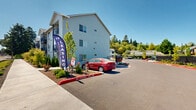 Trail Ave Apartments is a well-maintained residential community situated along Trail Ave NE in Keizer, Oregon. The property features multiple multi-story buildings finished in a clean light-blue and gray lap siding exterior, giving the community a fresh, modern aesthetic that stands out against the lush Pacific Northwest backdrop of mature trees and open skies.

The grounds are thoughtfully designed with newly landscaped planting beds, manicured shrubs, and young trees that line both the building perimeters and the sidewalk along Trail Ave NE. Clearly marked fire lanes and wide internal driveways ensure easy navigation throughout the property. Residents benefit from generous on-site parking, including multiple covered carport structures with sturdy metal canopies that provide weather protection across numerous spaces.

The community is secured with perimeter fencing and gated access points, contributing to a safe and private living environment. A dedicated leasing office on-site makes the rental process straightforward and accessible for prospective residents. Each building features exterior stairways and covered balconies or entry areas, adding both functionality and architectural interest. The overall atmosphere is clean, orderly, and move-in ready, making Trail Ave Apartments a practical and attractive residential option in Keizer.