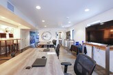 The Nest sales and leasing center is a thoughtfully designed, single-floor space that blends modern industrial style with warm, inviting elements to create an engaging environment for prospective residents. The layout flows naturally from a reception and display area—featuring bold blue cabinetry with sleek hardware, a wood-planked accent wall, and a community site map—into a series of functional yet comfortable zones.

The lounge area anchors the space with a rich leather sofa, a rustic wood coffee table, and an eclectic woven pendant light overhead. Natural light filters in through large windows framing outdoor greenery, while wall-mounted planter boxes and layered area rugs add texture and warmth. A teal accent wall with industrial pipe shelving provides visual contrast and showcases branded merchandise.

Adjacent to the lounge, a collaborative workspace features long communal tables with ample seating, ideal for meetings or one-on-one consultations. The space transitions into a dedicated presentation area equipped with individual computer workstations set against a textured accent wall with sculptural lighting fixtures, providing prospective residents with an interactive way to explore floor plans and community details.

Throughout, warm hardwood-style flooring, recessed lighting, and carefully curated décor maintain a cohesive aesthetic that is both professional and approachable, reflecting the community's "Living the Nest Life" brand identity.