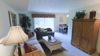 This single-floor residence offers a comfortable and classic interior design, centered around a bright, open-concept living and dining area. The living room is warmly appointed with plush, cushioned sofas accented by rich burgundy and earth-tone throw pillows, arranged around a charming white cottage-style coffee table resting atop a decorative area rug. A large window dressed with an elegant floral valance and vertical blinds allows abundant natural light to fill the space, creating an airy and welcoming atmosphere. Lush indoor plants add a touch of greenery and life to the corners of the room, complemented by a stately pine armoire that brings a classic, rustic character to the space. The living area flows seamlessly into the dining area, which features a dark wood dining table and matching chairs, illuminated by a ceiling fan with a light fixture overhead. Framed artwork on the walls adds a personalized touch throughout. The neutral-toned carpet throughout the space ties the rooms together cohesively, contributing to the overall warmth and comfort of this well-curated home.