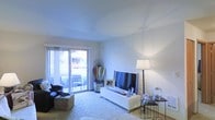 This single-floor space offers a welcoming and well-lit environment designed for comfortable everyday living. The living area serves as the heart of the space, featuring smooth white walls and a clean, neutral ceiling that enhances the sense of openness and airiness throughout. A large sliding glass door floods the room with abundant natural light and provides direct access to an outdoor patio area, where seating and greenery are visible, offering a seamless indoor-outdoor connection. The living room is thoughtfully arranged with a plush area rug anchoring the seating area, a modern white media console supporting a flat-screen television, and an elegant floor lamp that adds warm accent lighting. Decorative touches — including framed artwork, glass accents, and a vintage wooden crate used as a side table — lend the space a charming, personalized character. A tall side table with glass and metallic accessories adds a refined finishing touch to the corner of the room. The overall ambiance is light, airy, and inviting, with a casual yet curated aesthetic that balances functionality and style throughout.